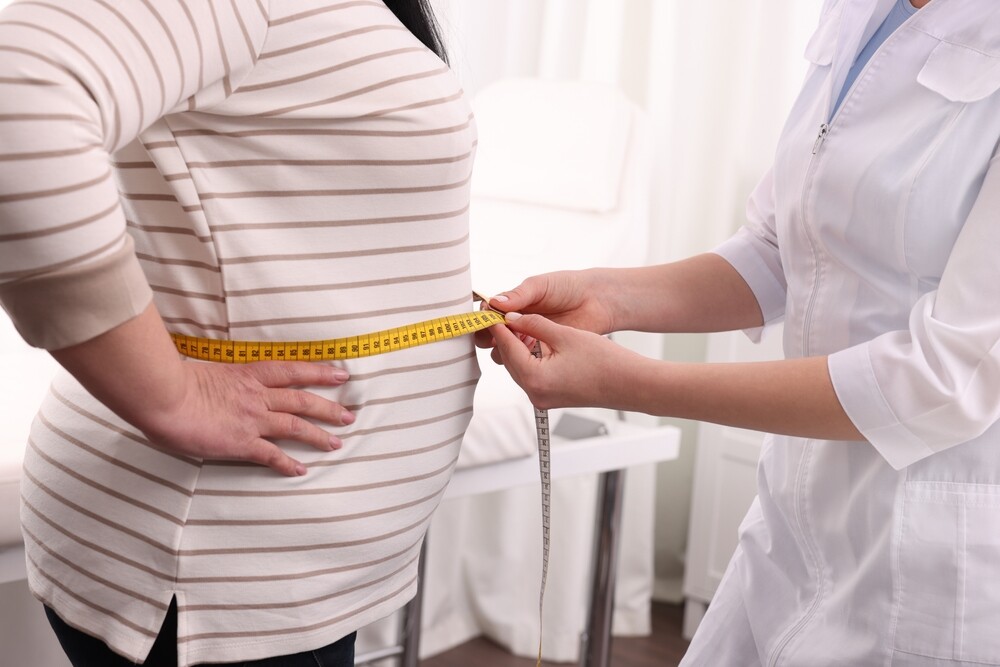 Nutritionist measuring overweight woman's waist with tape in clinic, closeup.