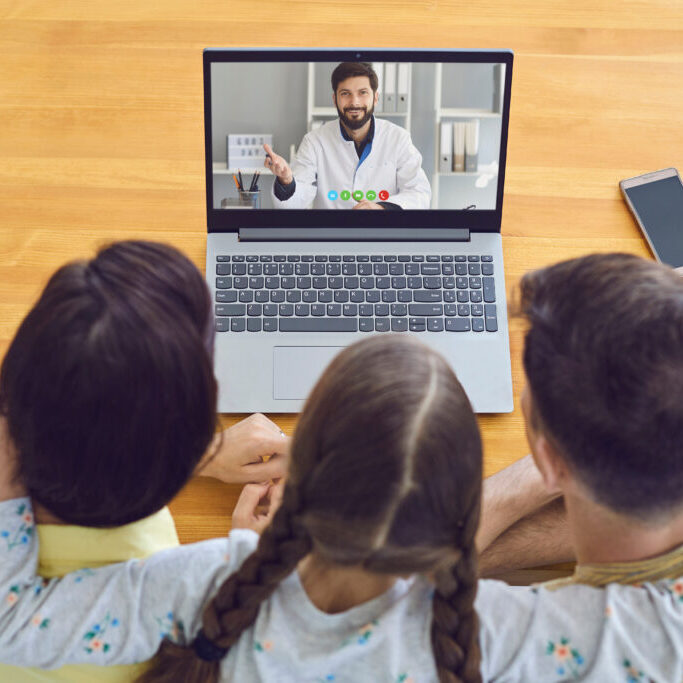 Family sitting in front of a laptop video chatting with their doctor.
